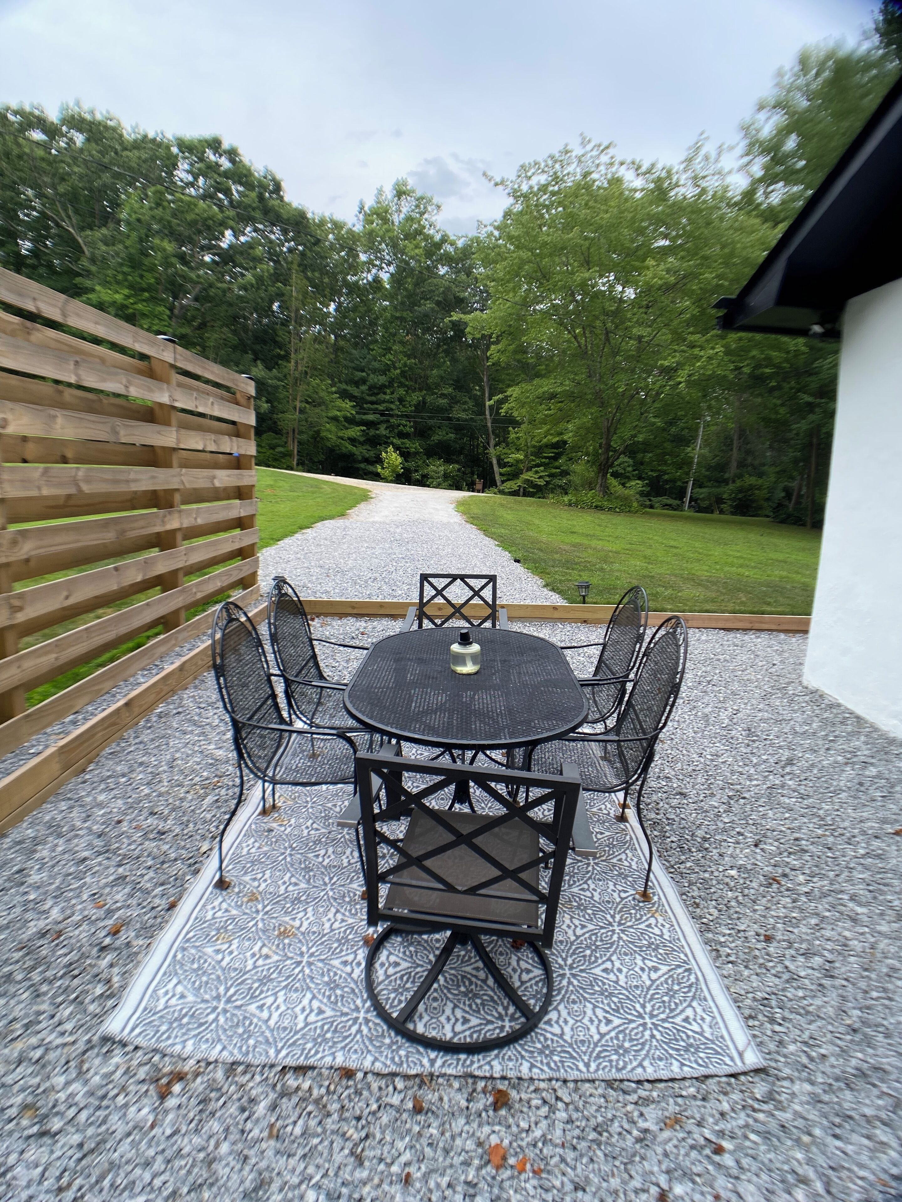 A patio with a round table and four chairs, surrounded by a wooden fence and a gravel path leading to a grassy area.