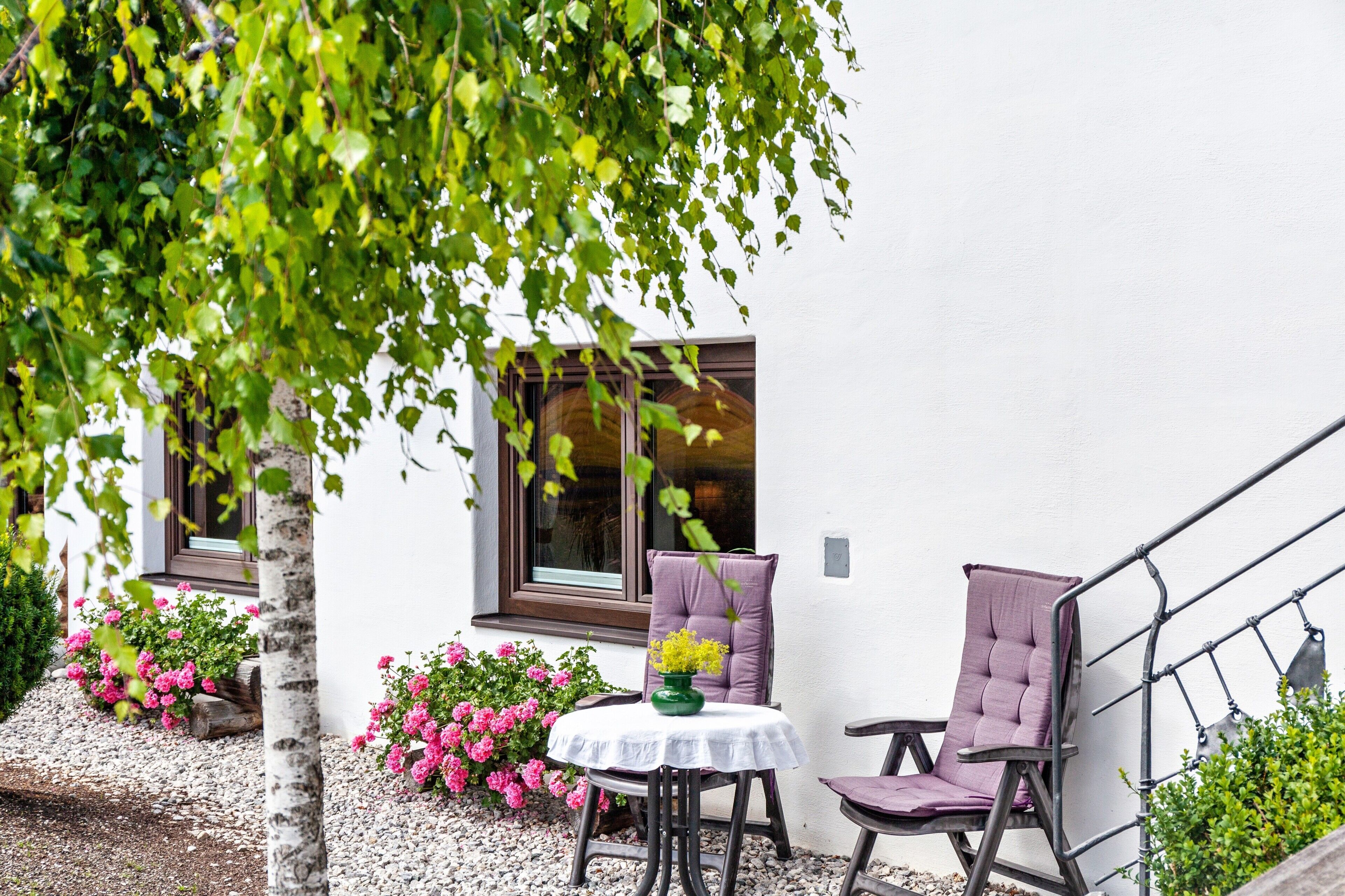En terrasse med et bord og to stole, omgivet af en hvid bygning med vinduer og et træ.