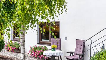 En terrasse med et bord og to stole, omgivet af en hvid bygning med vinduer og et træ.