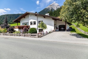 A white building with a wooden roof, a paved driveway, and a mountain in the background.