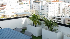 En balkon med et bord og stole, omgivet af hvide plantekasser med grønne planter, med udsigt over et bybillede med hvide bygninger.