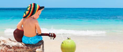 A child sitting on a wooden log at the beach, holding a toy guitar, with a green coconut in front of them.