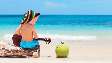 Niño sentado sobre un tronco de madera en la playa, sosteniendo una guitarra de juguete, con un coco verde frente a él.