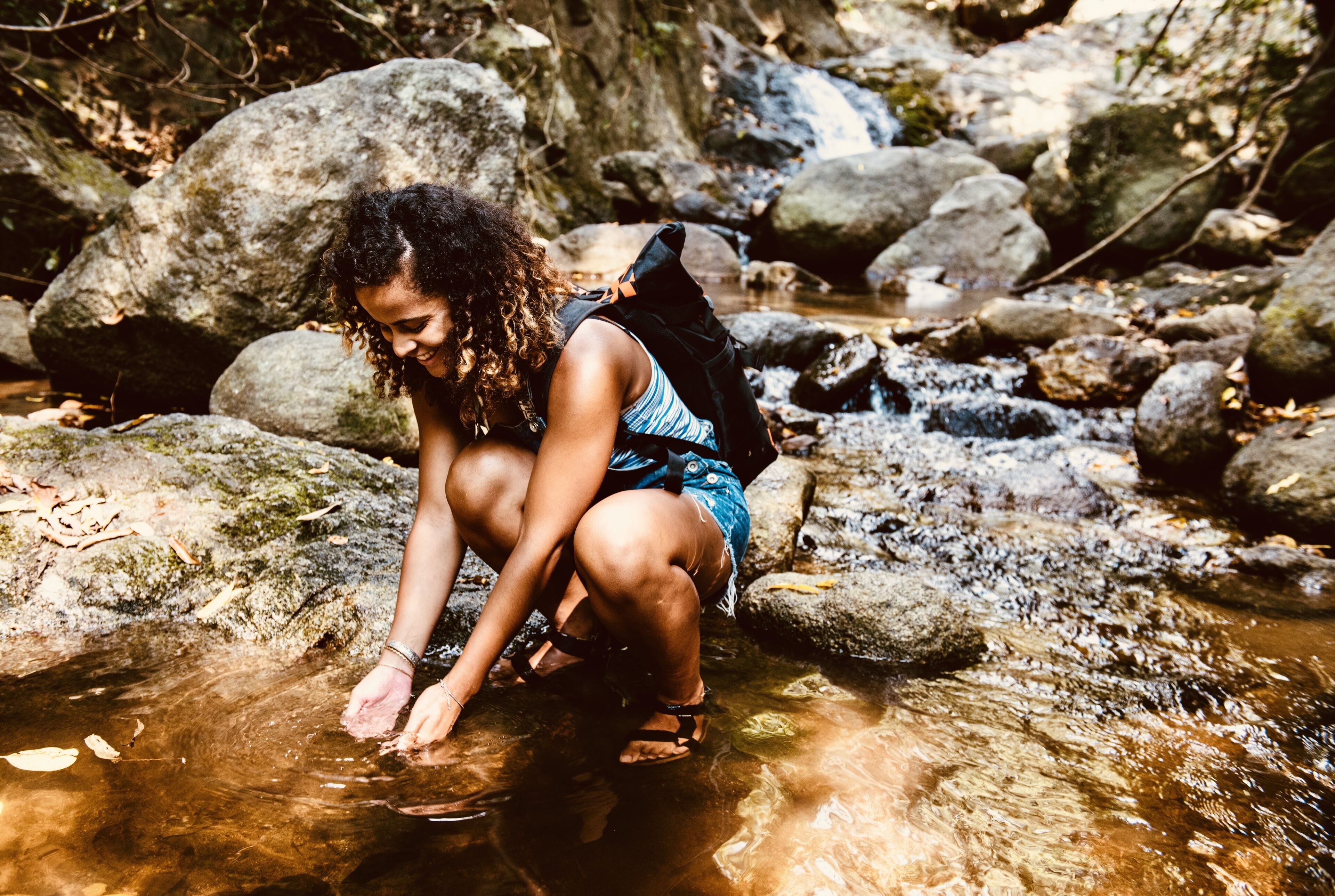 A person crouching by a clear stream surrounded by large rocks and foliage.