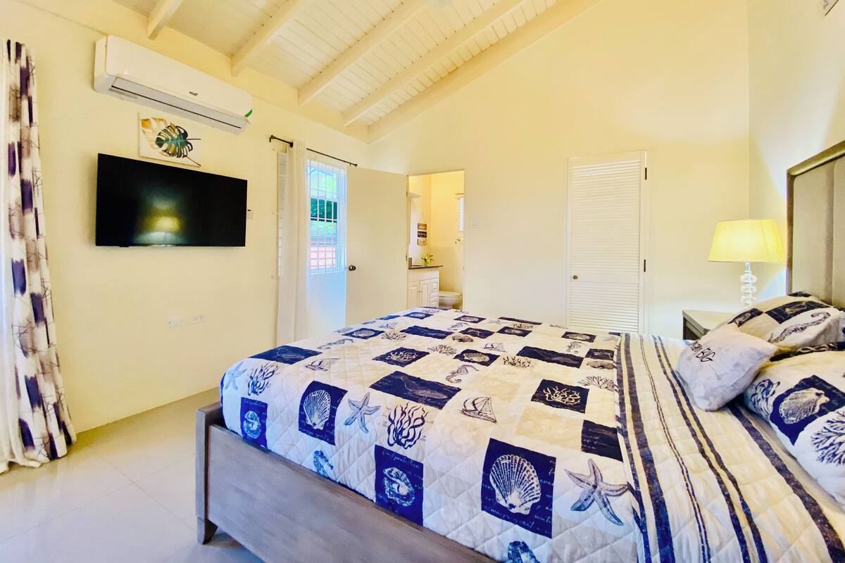 Ein Schlafzimmer mit Bett, Fernseher, Klimaanlage und einem durchgehenden Türbogen sichtbarem Badezimmer.