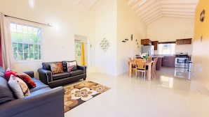 Een woonkamer met een zwarte leren bank, een eettafel met stoelen, een keuken met een fornuis en een koelkast, en een groot raam met gordijnen.