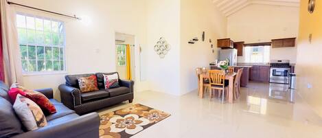 Una sala de estar con un sofá de cuero negro, una mesa de comedor con sillas, una zona de cocina con estufa y refrigerador, y un ventanal con cortinas.