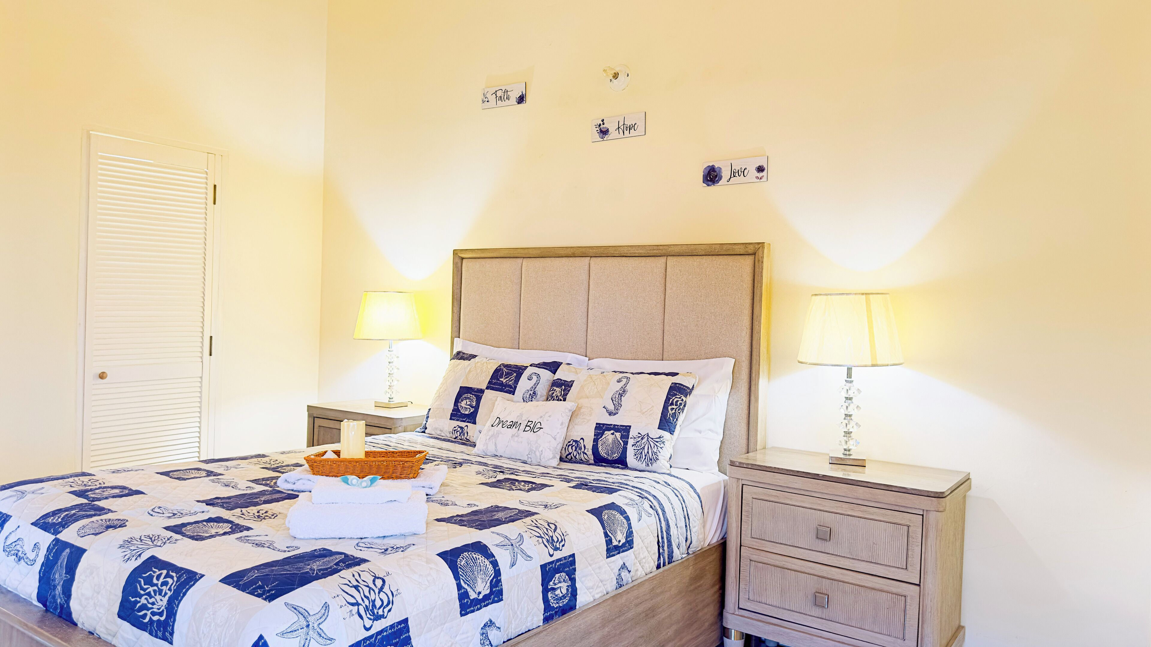 A neatly made bed with a blue and white quilt, two bedside lamps, a wooden nightstand, and a wall with inspirational signs.