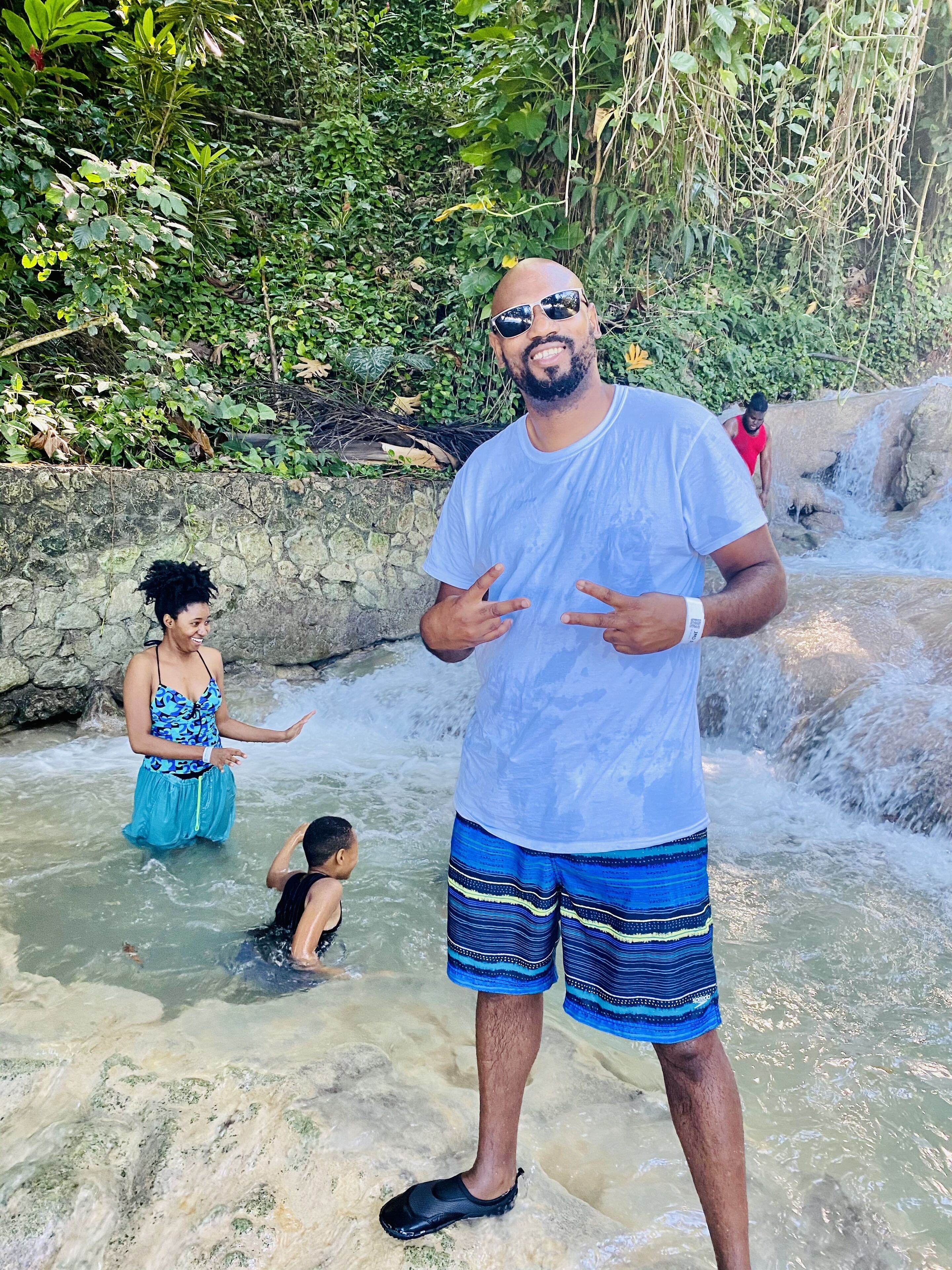 A man standing in a shallow water area with a rocky wall and greenery in the background.