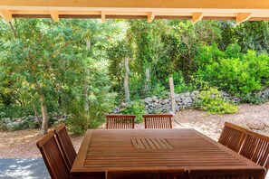 Une terrasse extérieure en bois, avec une table et quatre chaises, entourée d’une végétation luxuriante et d’un mur de pierre.