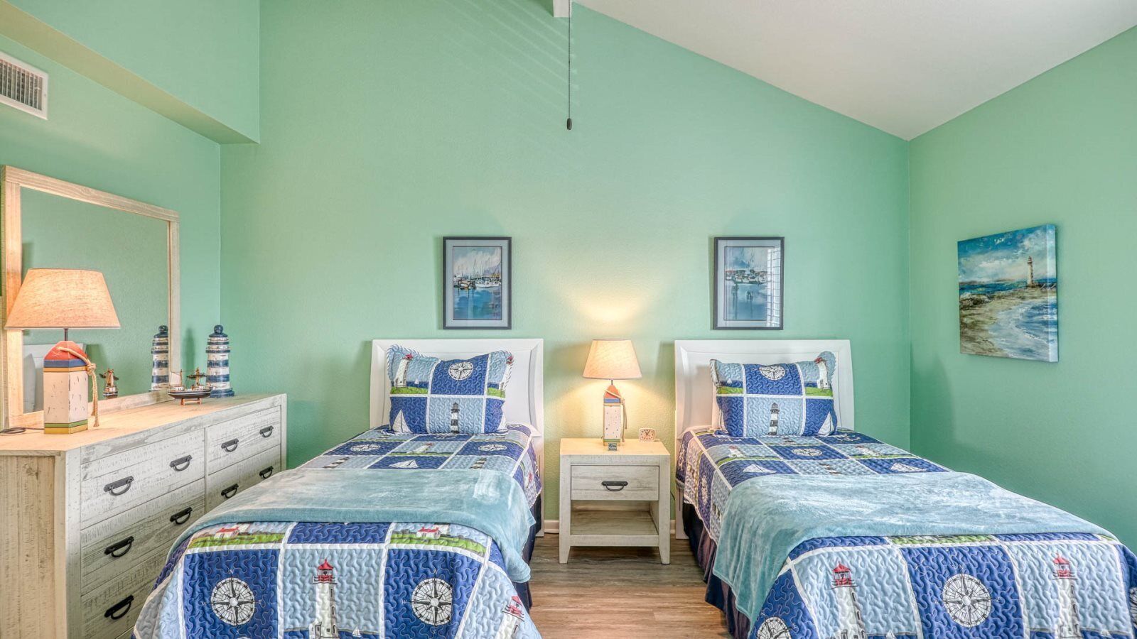 Ein Zimmer mit zwei Betten, jeweils mit einer blau-weißen Steudecke, einem Nachttisch mit Lampe, einem Schminktisch mit Spiegel und einem gerahmten Bild an der Wand.