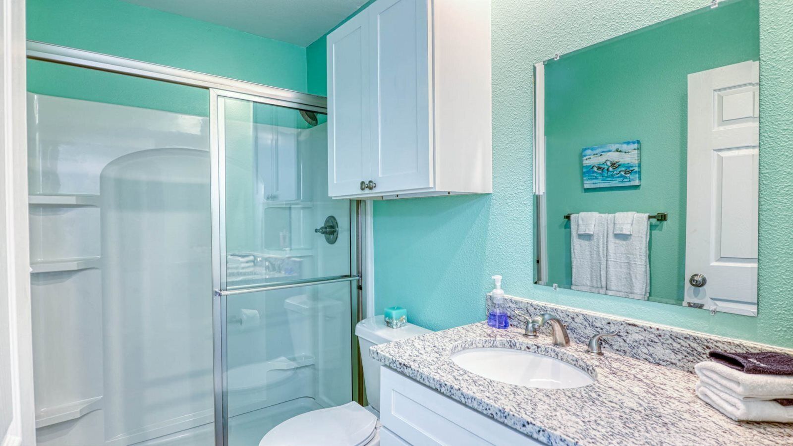A bathroom with a glass-enclosed shower, a white toilet, a sink with a marble countertop, and a mirror with a towel rack.