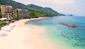 Una playa con agua turquesa cristalina, una orilla arenosa y un hotel de varios pisos con balcones con vistas a la playa.