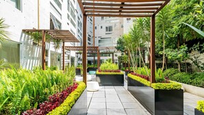 A modern outdoor corridor with a pathway lined by lush greenery and black planters, leading to a building with multiple floors and large windows. - Most amazing view of Mexico City. Pool + gym + parking + AC (Ciudad de México)