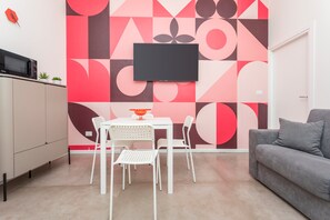 A modern living room with a flat-screen TV mounted on a geometric-patterned wall, a grey sofa, and a white dining table with chairs. - Taormina Center Suite Apartment with In-Room Jacuzzi (Taormina)