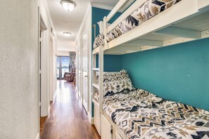 A bunk bed with patterned bedding, a hallway with wooden flooring, and a view into another room with a chair.