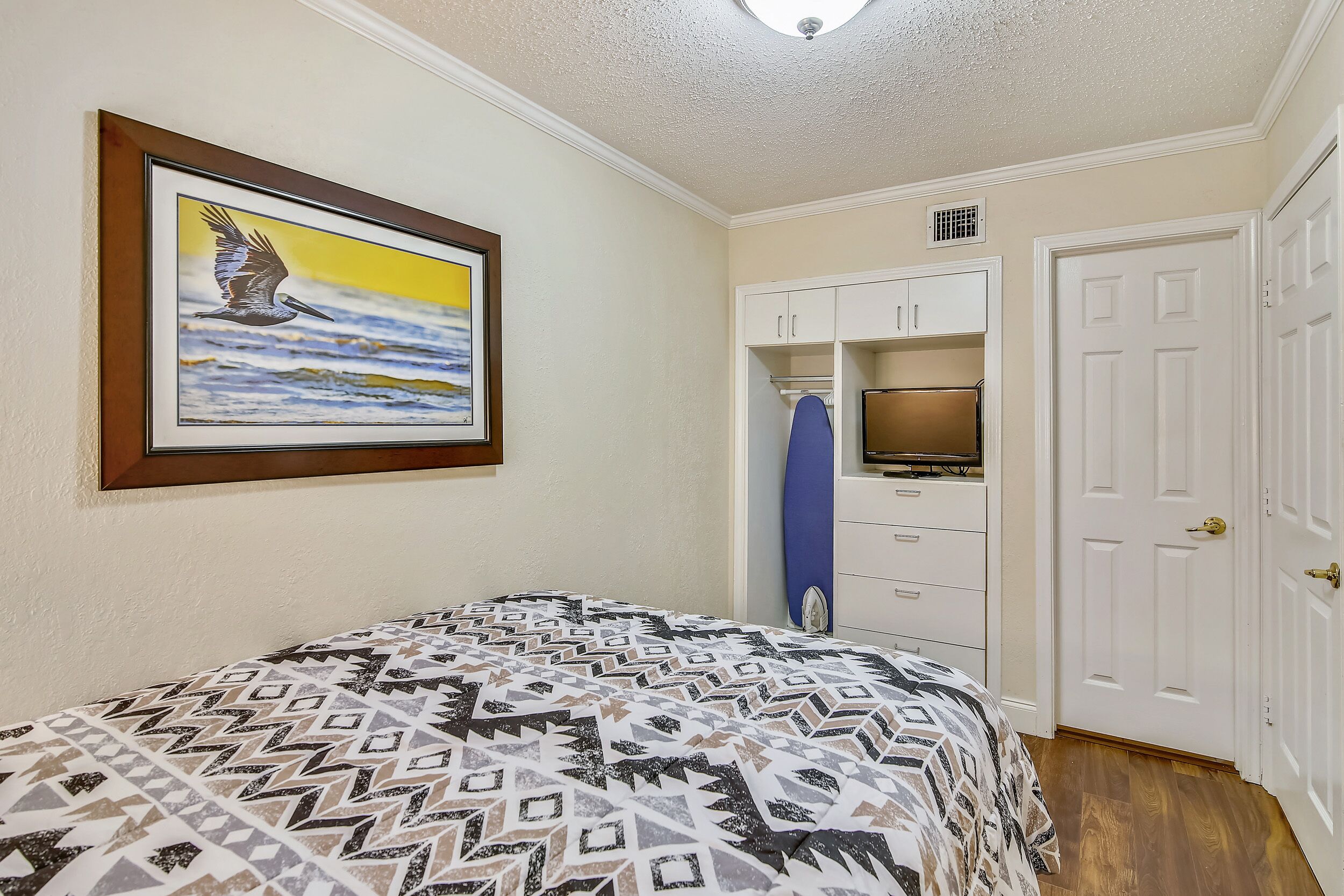 A bedroom with a bed, a framed picture, a kitchenette, and a white door.