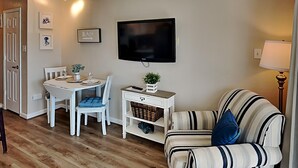 A living room with a TV, a striped sofa, a dining table with chairs, and a small side table with a plant. - Casa Del Mar by Ryson Vacation Rentals (Galveston)