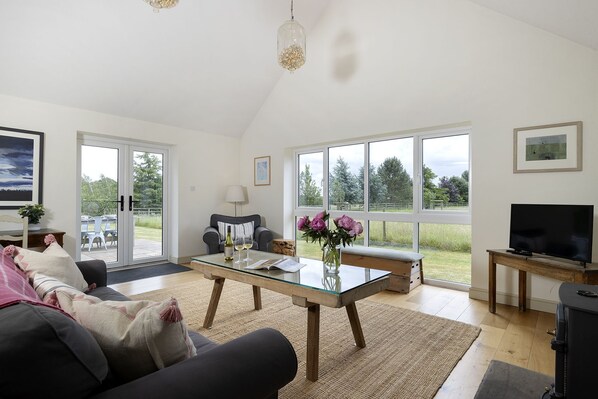 A spacious living room with a glass coffee table, a sofa, a television, and large windows offering a view of the outdoors.