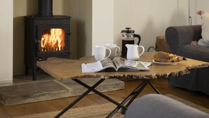 A cozy living room with a wood-burning stove, a coffee pot, a plate of bread, and a book on a rustic table. - Pet Friendly Rural Cotswold Cottage - 1 Manor Farm Cottage (Hook Norton, Banbury)