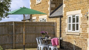 A patio with a table, chairs, a green umbrella, and a wooden fence. - Pet Friendly Rural Cotswold Cottage - 1 Manor Farm Cottage (Hook Norton, Banbury)