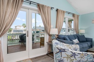 Una sala de estar con un sofá azul, cortinas color beige y vista a una casa y palmeras a través de un ventanal.