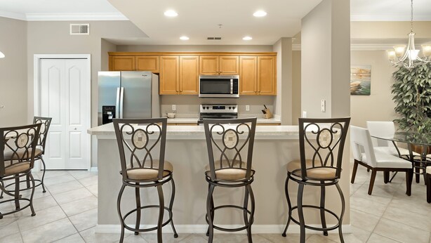 A modern kitchen with wooden cabinets, a stainless steel refrigerator, a microwave, and a built-in oven. There are three bar stools with cushioned seats and a glass-top dining table with chairs. - Riverwalk 713 by Distinctive Beach Rentals (Fort Myers)
