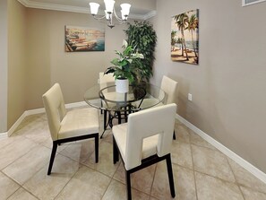 A dining area with a glass table surrounded by four chairs, a potted plant, and two paintings on the walls. - Riverwalk 713 by Distinctive Beach Rentals (Fort Myers)