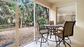 A covered patio with a table and chairs, a view of a natural landscape through large glass doors, and a window with blinds. - Riverwalk 713 by Distinctive Beach Rentals (Fort Myers)