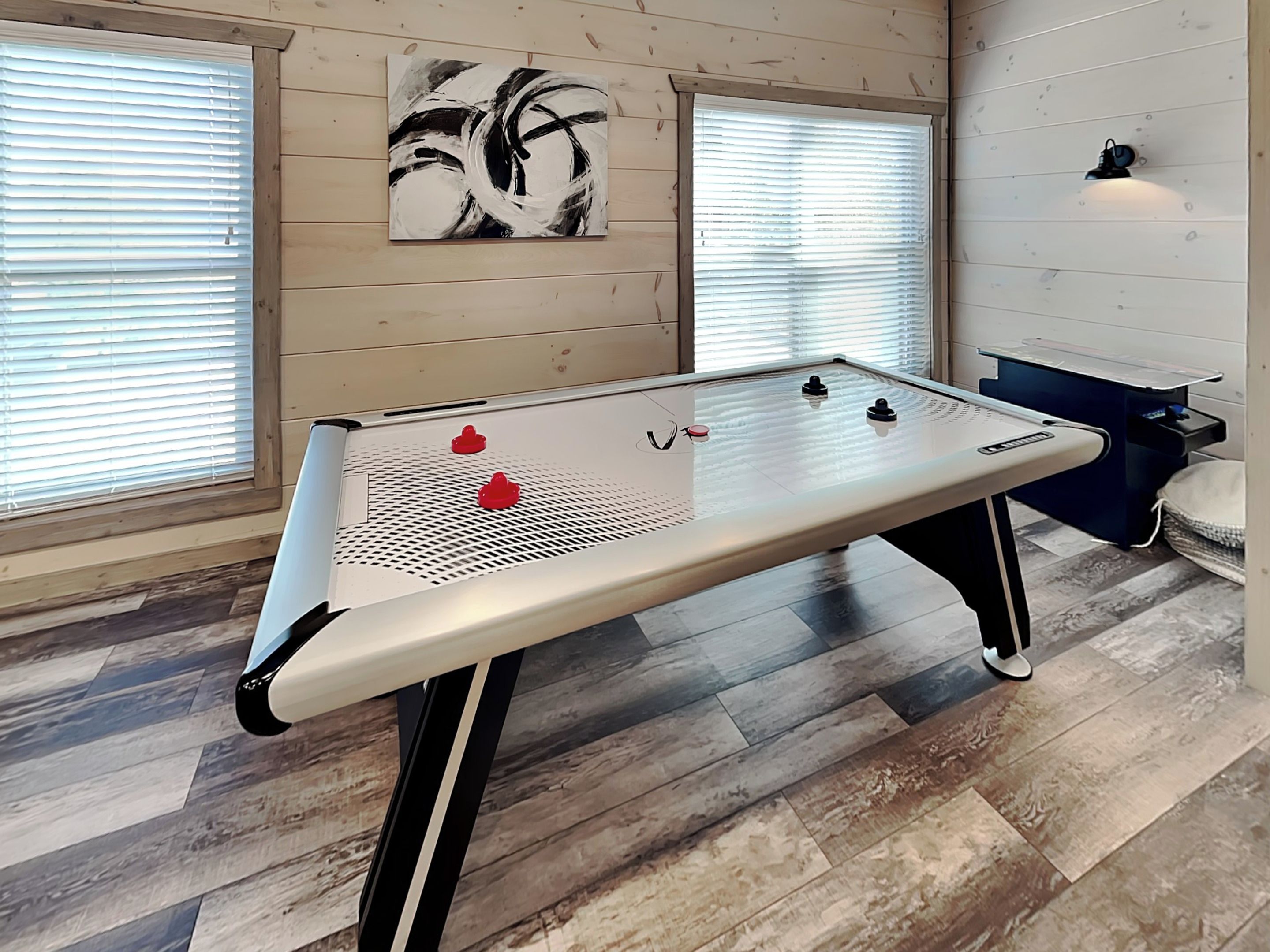 A foosball table in a room with wooden floors and walls, a painting on the wall, and a window with blinds.