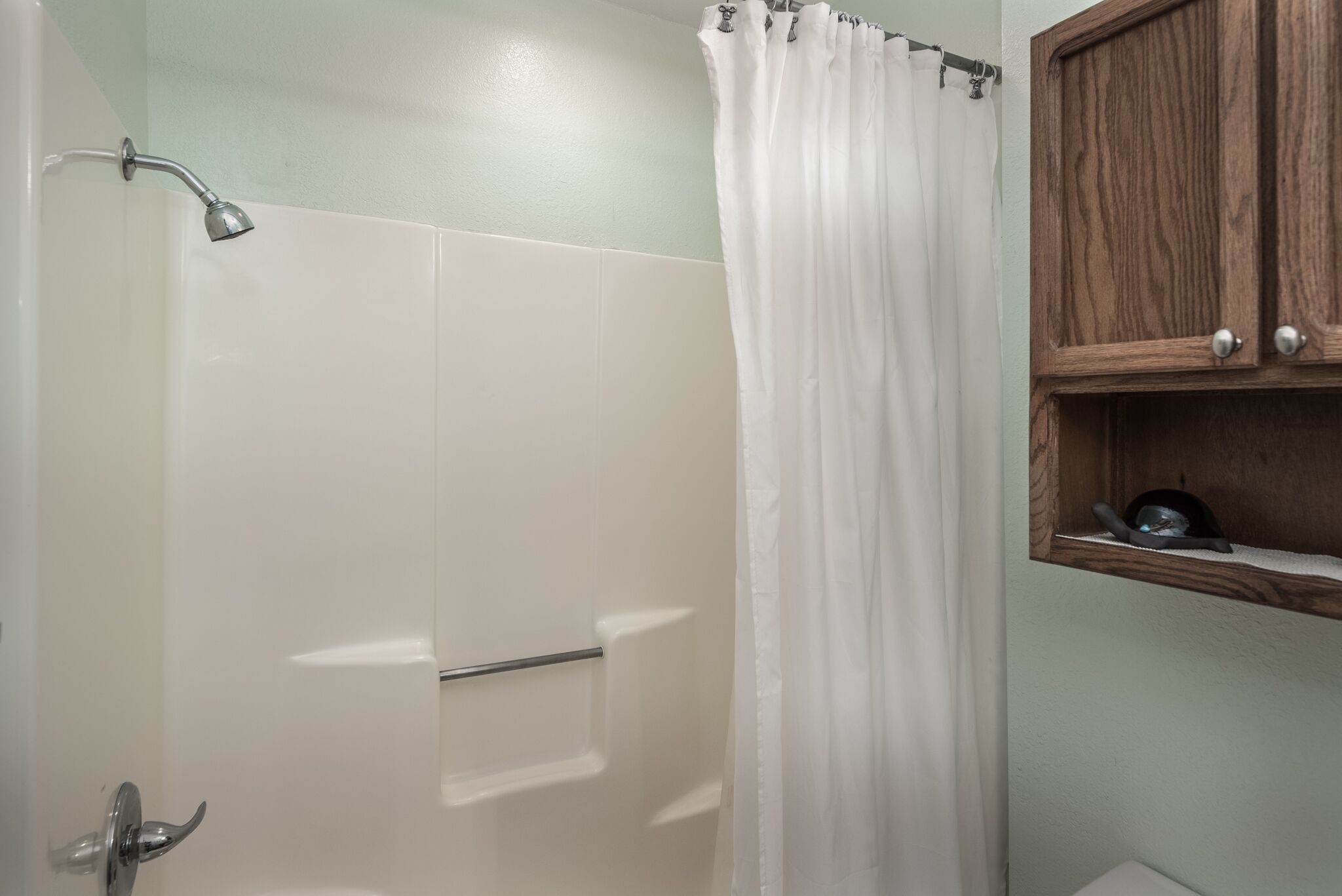 A bathroom with a shower, a white curtain, and a wooden shelf with a pan.
