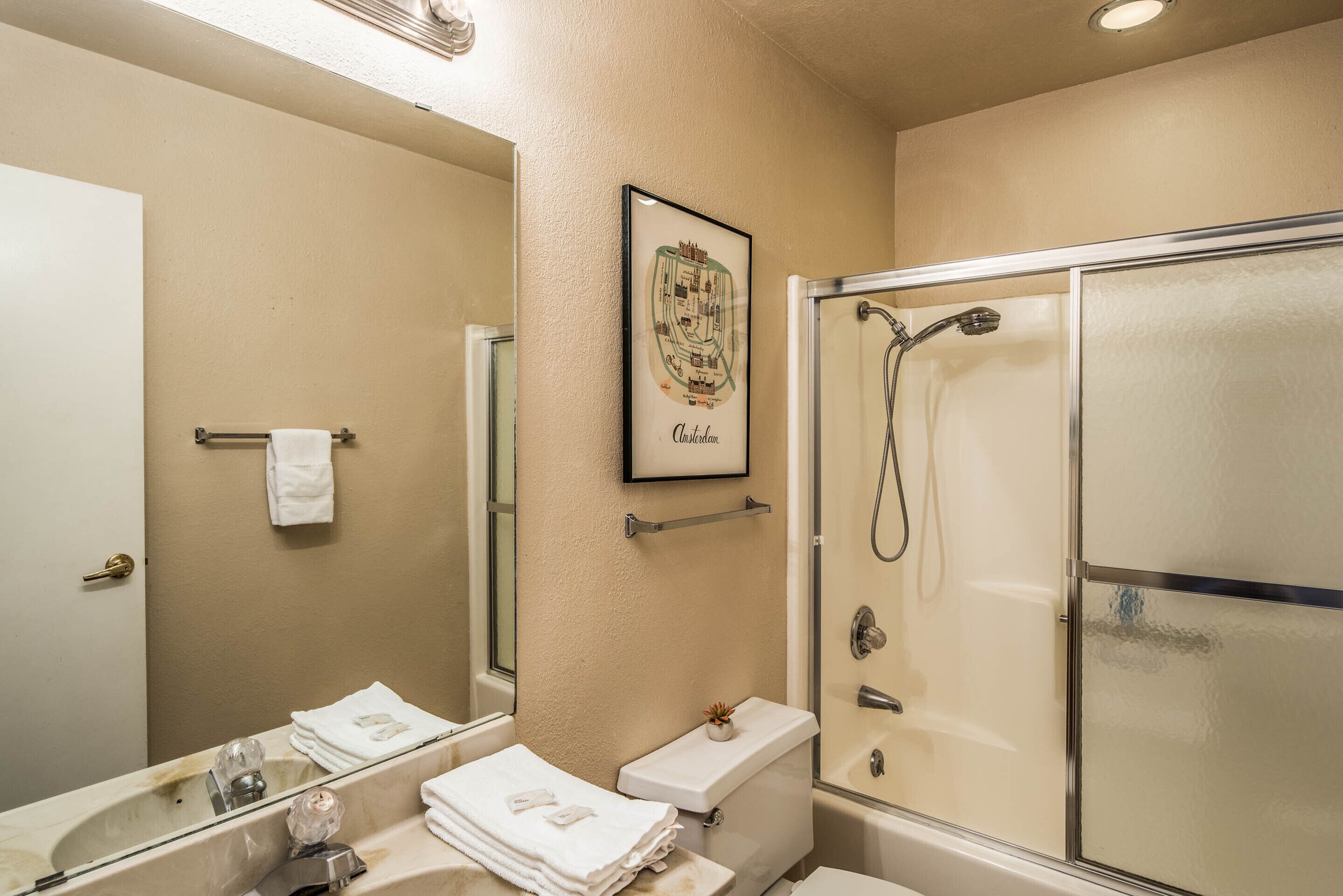 A bathroom with a glass-enclosed shower, a sink with a mirror, and a framed picture on the wall.