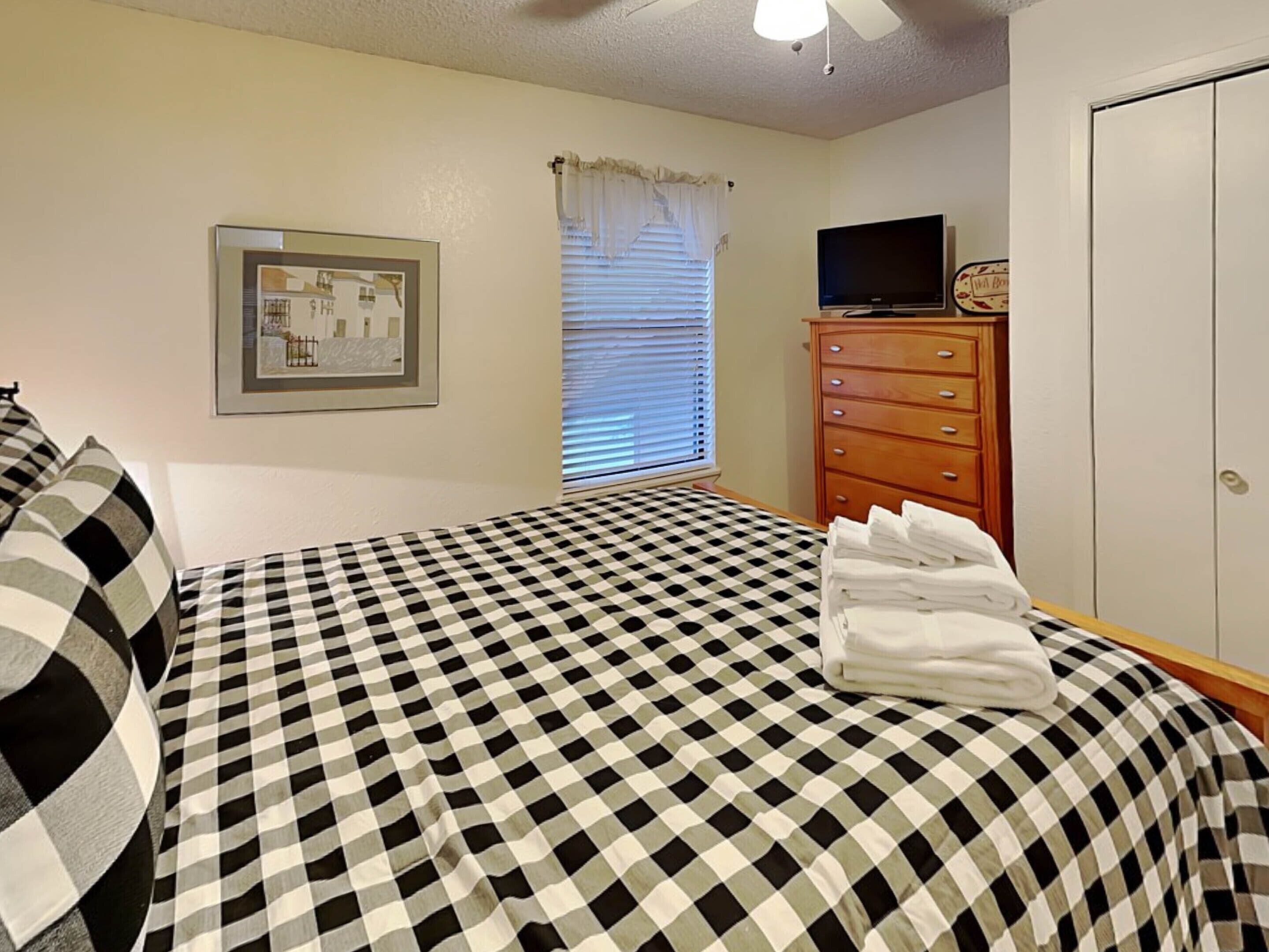 A bedroom with a checkered bedspread, a framed picture on the wall, a television on a wooden dresser, and a window with blinds.