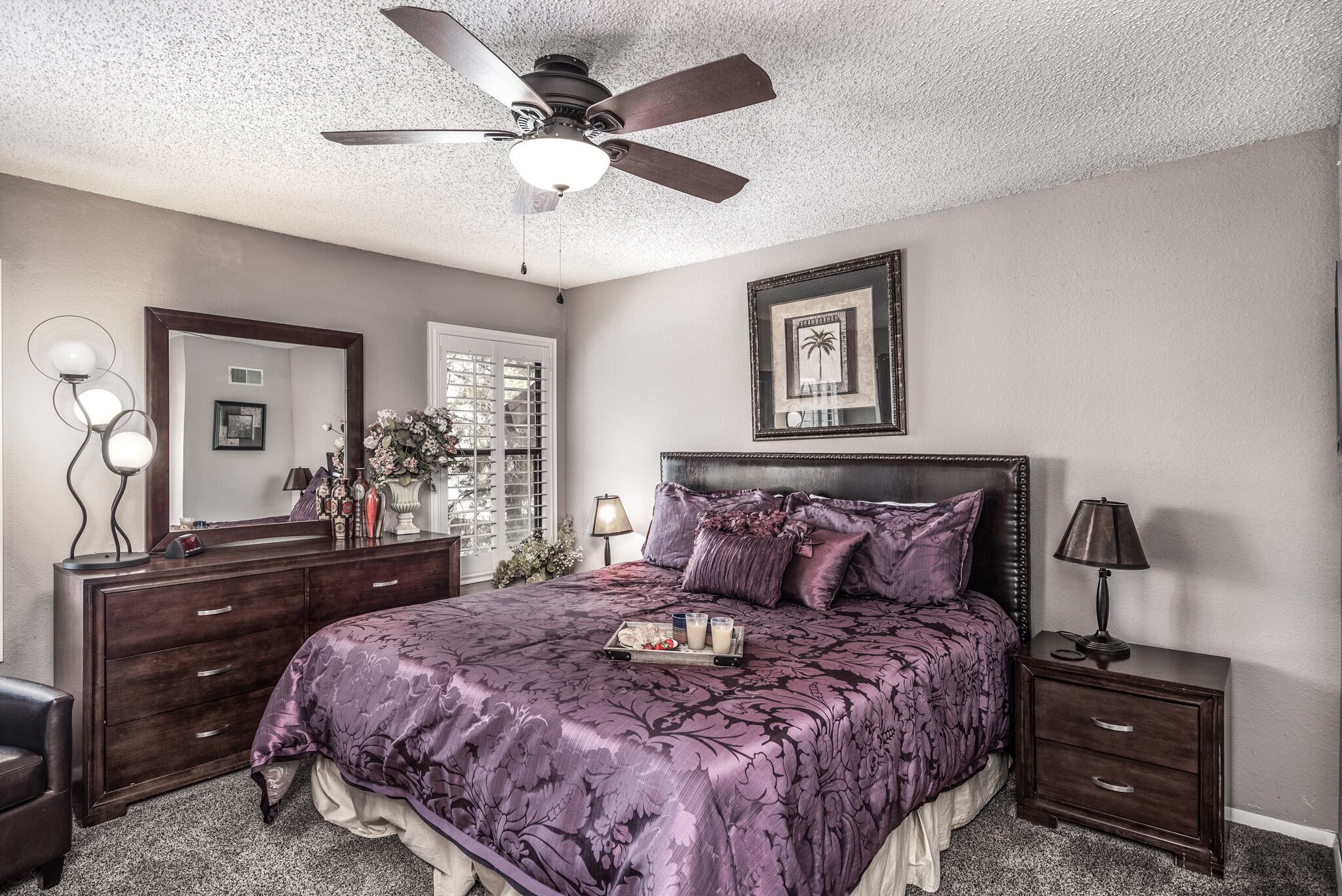 A bedroom with a large bed, a ceiling fan, a dresser, a nightstand, and a framed picture on the wall.