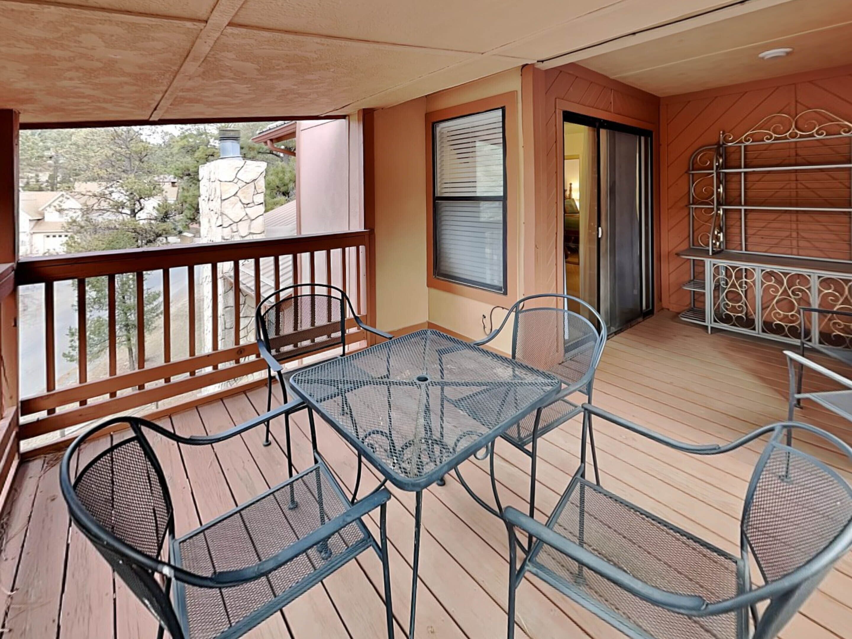 Un patio techado al aire libre con piso de madera, mesa y sillas de metal, baranda y vista del entorno.
