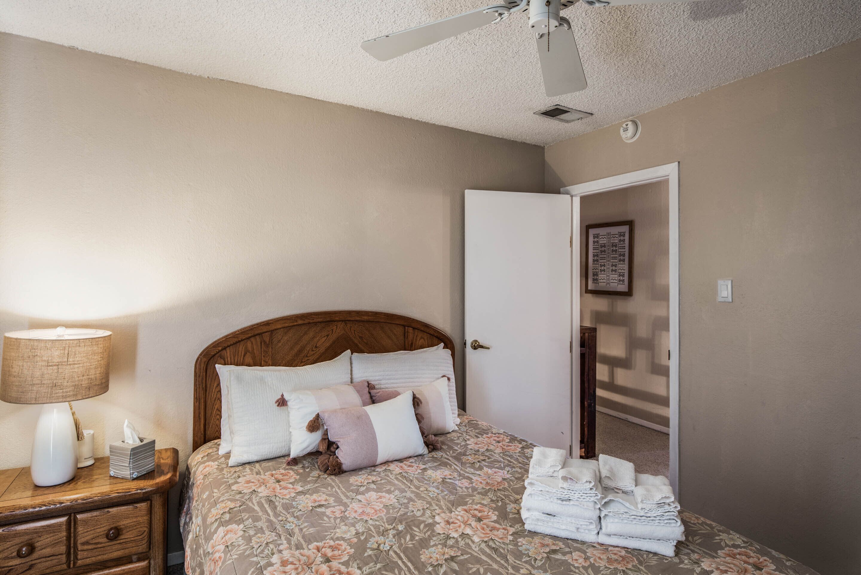 Un dormitorio con cabecera de madera, cama con ropa de cama floral, mesita de noche con lámpara y una puerta con un cuadro enmarcado.