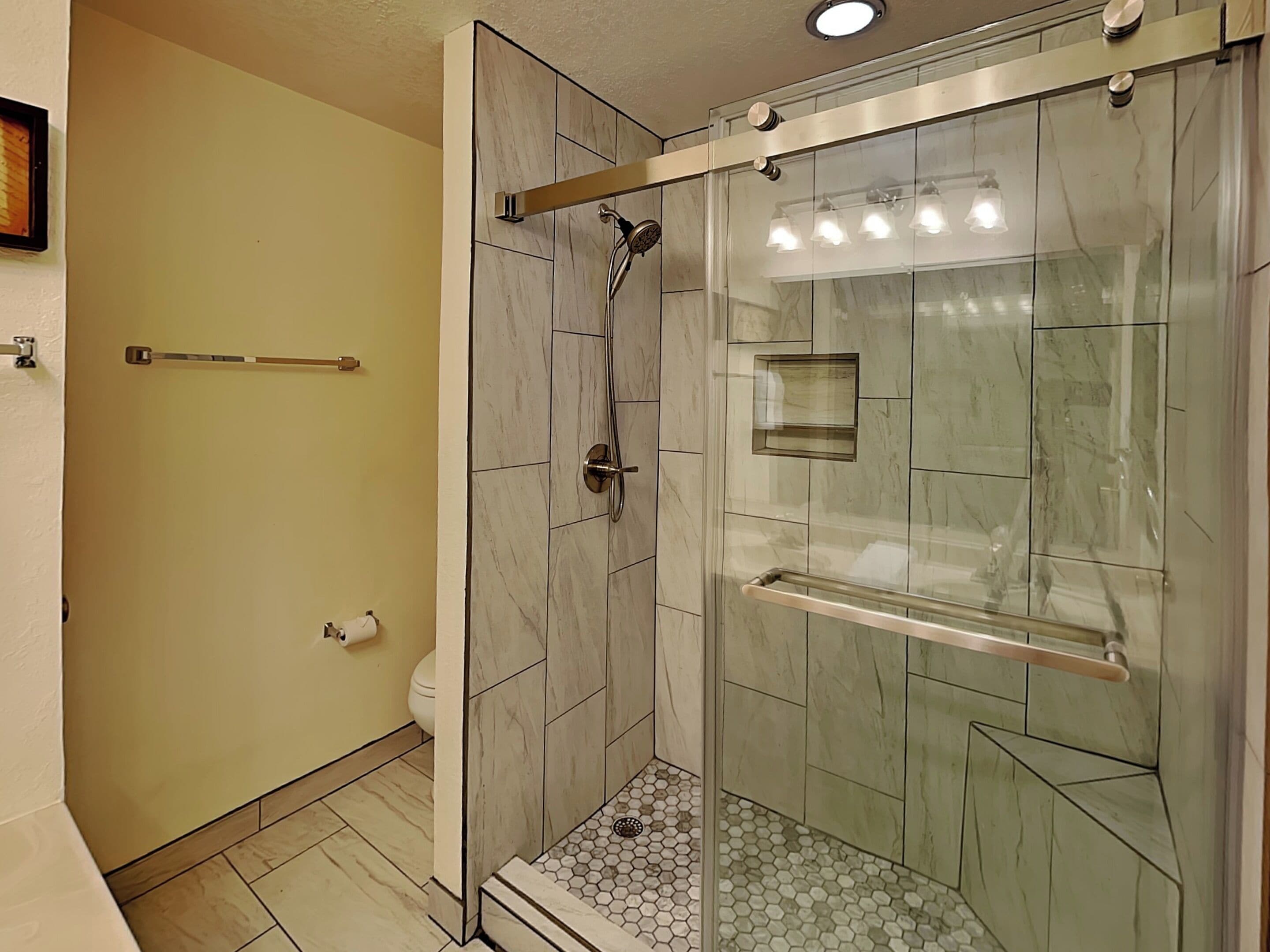 A glass-enclosed shower with a handheld showerhead, a glass shelf, and a toilet with a toilet paper holder.