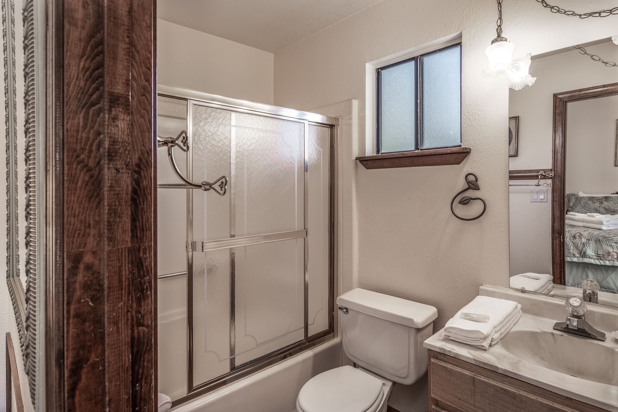 Une salle de bain avec une douche à parois de verre, des toilettes blanches, un lavabo avec un miroir et une fenêtre située au-dessus des toilettes.
