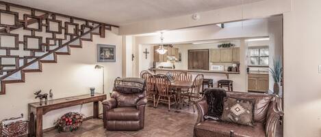 A spacious living area with a wooden staircase, leather furniture, a dining table, and a kitchen in the background.