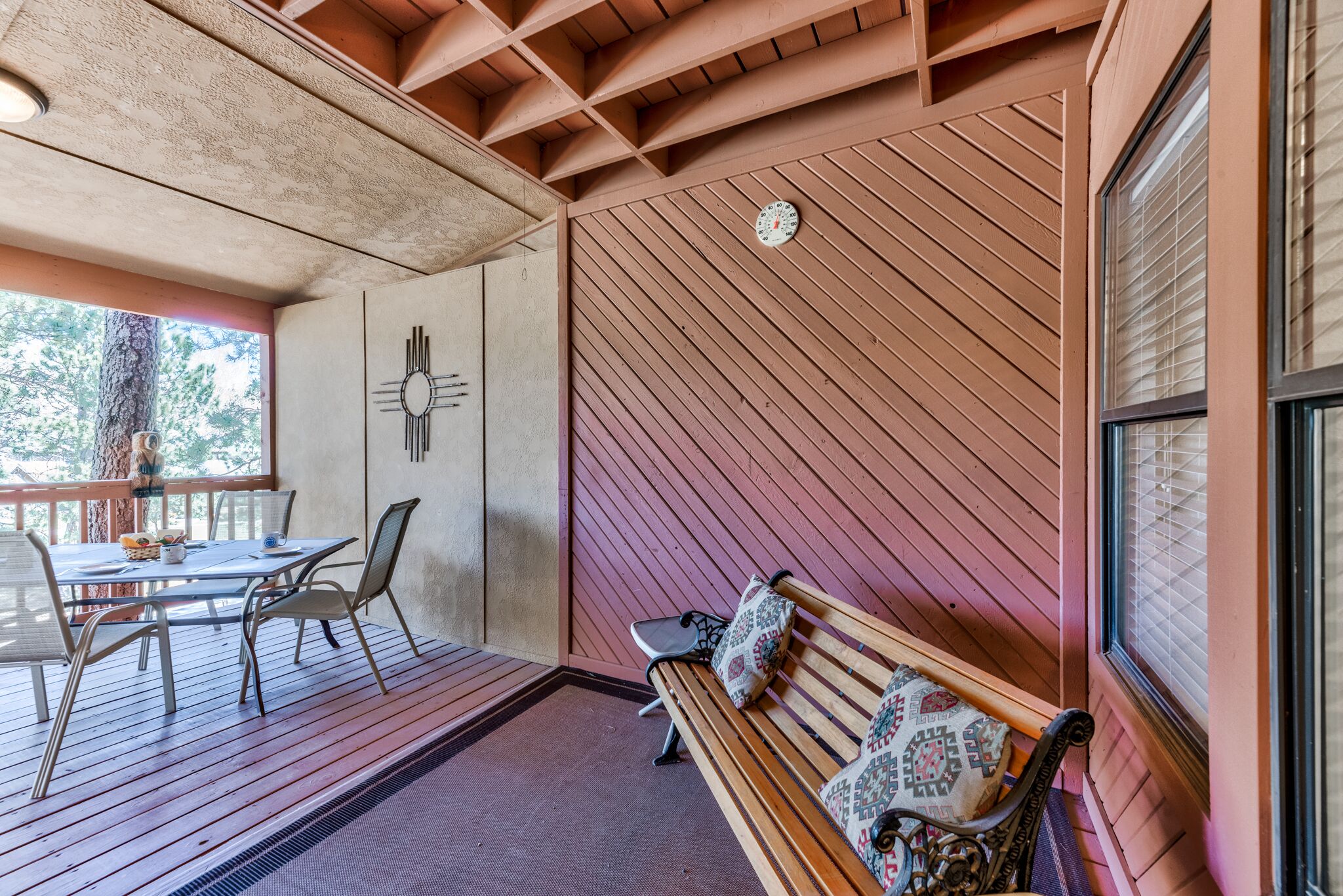 A wooden deck with a table and chairs, a bench with patterned cushions, and a wall clock.