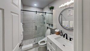 A modern bathroom with a glass-enclosed shower, a toilet, a sink with a marble countertop, and a mirror with a round frame.