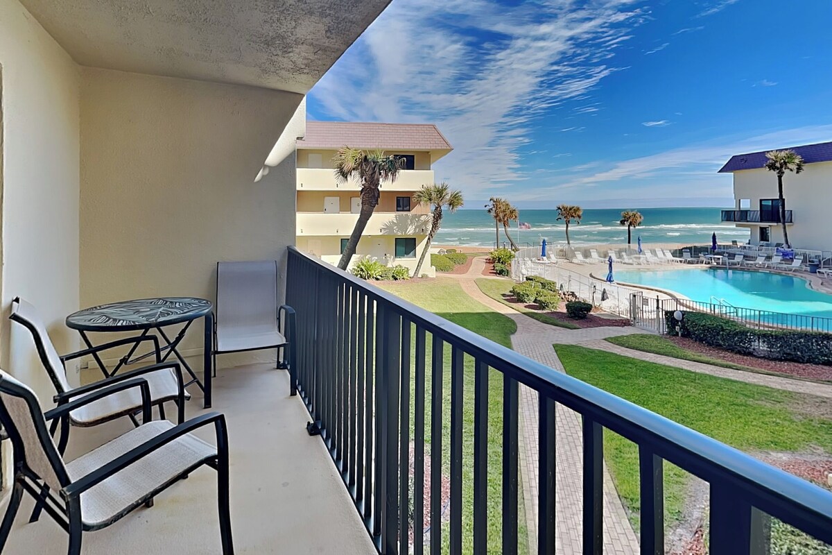 A balcony with a pool view, outdoor seating, and a clear sky.