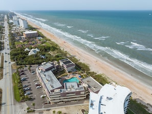 An aerial view of a coastal hotel with a swimming pool, beach access, and adjacent high-rise buildings.