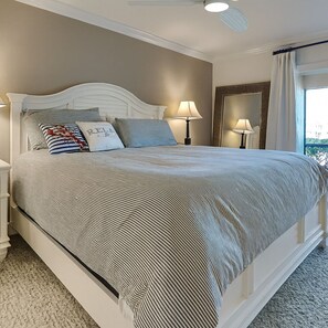A bedroom with a large bed, two bedside lamps, a ceiling fan, and a window with curtains.