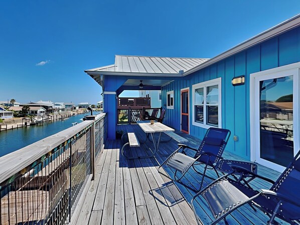 Un edificio azul con terraza de madera, asientos exteriores y vista a un puerto deportivo.