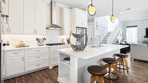 A modern kitchen with white cabinetry, a large island with stools, a hanging light fixture, and a staircase leading to a living area. - Key West by Ryson Vacation Rentals (Galveston)