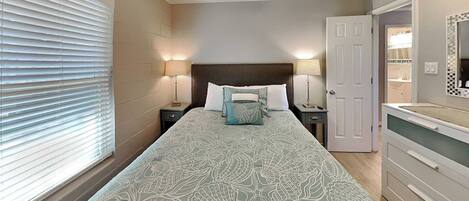 A bedroom with a bed, two bedside lamps, a ceiling fan, a window with blinds, and a door leading to a bathroom.