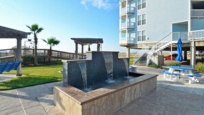 A pool area with a water feature, picnic tables, and a building with balconies. - Abundant Relaxation by Ryson Vacation Rentals (Galveston)