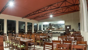 A restaurant with multiple dining tables and chairs, a kitchen area, and large windows. - The Tattwaa Corbett Spa & Resort (Ramnagar)