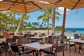 An outdoor dining area with wicker chairs and tables, overlooking a beach and ocean.